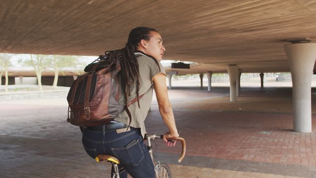 A man with long dreadlocks rides a bicycle under an urban bridge on a sunny day. Perfect for use in promoting sustainable urban transport, city life exploration, or lifestyle brands targeting bikers and eco-conscious consumers.