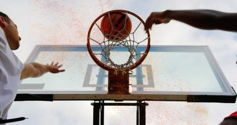 Underhoop action: basketball passing through rim as two players reaching, powder burst