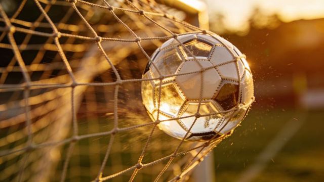 Backlit soccer ball hitting net at golden hour with water spray and goal impact