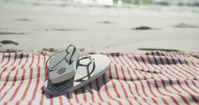White flip flops are placed on a red and white striped towel on the sandy beach near the ocean. Ideal for summer vacation promotions, travel advertisements, beach lifestyle content, and relaxation-themed marketing. This scene evokes a sense of leisure and comfort, making it suitable for use in travel brochures, summer campaigns, and social media posts.