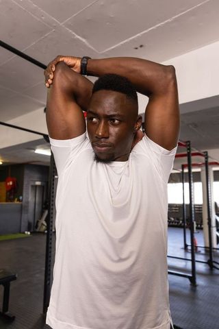 African american man stretching overhead by squat rack wearing smartwatch in gym