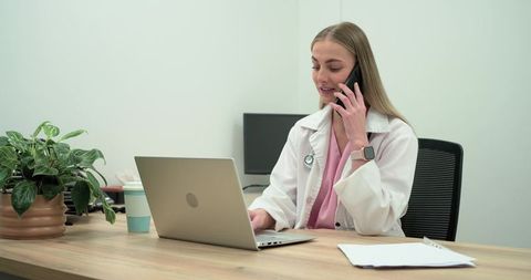 Female doctor talking on smartphone while typing on laptop in modern clinic workspace