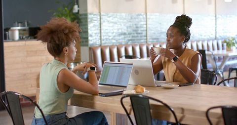 Two women collaborating on laptops in cafe coworking space, drinking coffee, planning work
