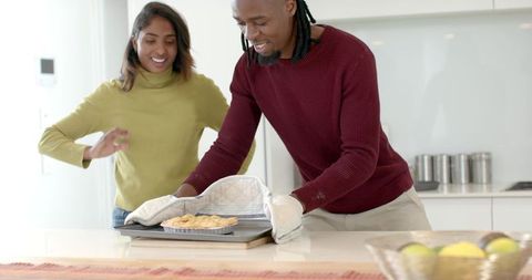 Smiling diverse couple removing freshly baked pie from oven at bright modern kitchen island