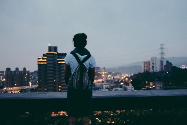 Silhouette of a person wearing a backpack standing on a rooftop, looking out at the cityscape during dusk. The person's contemplative stance and the twinkling city lights create a peaceful and introspective mood. Useful for themes like solitude, contemplation, urban life, travel, adventure, and city views.