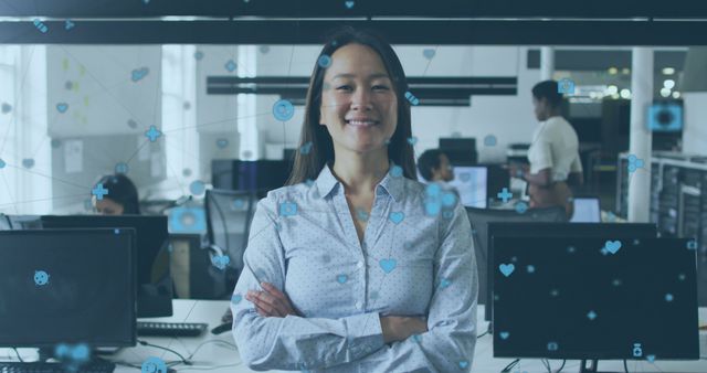 A confident businesswoman is standing with arms crossed in a modern tech office, smiling at the camera. The background includes colleagues working at their desks and multiple computer screens. Digital icons representing technology and innovation are overlaid on the image, adding a futuristic touch. This stock photo is ideal for business, technology, innovation, career, and professional success themes for use in websites, blogs, presentations, and corporate materials.