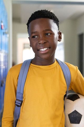 Smiling african american schoolboy holding soccer ball with backpack in school hallway