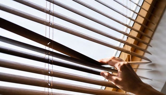 Hand adjusting wooden venetian blinds, sunlight filtering through slats
