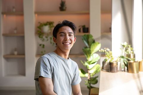 Young asian man smiling while sitting at sunlit wooden counter with indoor plants
