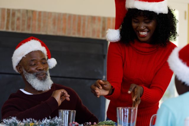 African American family members are celebrating Christmas dinner together, wearing Santa hats and sharing joyful moments. This image can be used for holiday greeting cards, festive advertisements, family-oriented promotions, or articles about holiday traditions and family gatherings.
