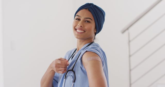 Nurse wearing blue scrubs with a headscarf, holding her sleeve up to show a bandage on her upper arm, signifying she has received a vaccination. The nurse is smiling, creating a positive and reassuring atmosphere. This image can be used to promote vaccination, healthcare awareness, medical services, and the importance of healthcare professionals. Ideal for hospital, clinic, and medical organization communications.