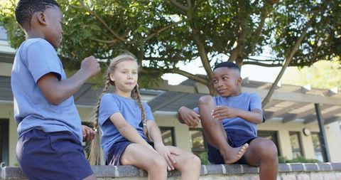 Schoolyard kids sitting on low brick wall under tree wearing blue uniforms casual playtime