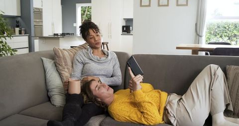 Two women relaxing on couch browsing tablet and holding mug in cozy open-plan living room