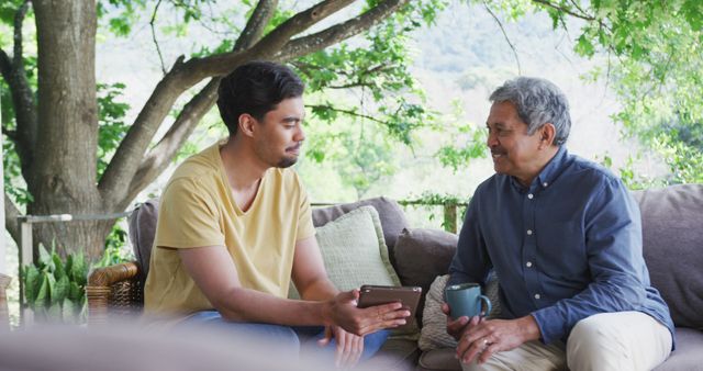 A serene moment between an elderly father and his adult son enjoying a conversation on an outdoor sofa. This image captures intergenerational bonding and familial connections, ideal for use in contexts such as family-oriented advertisements, retirement living promotions, Father's Day campaigns, and articles about family relationships or home life.