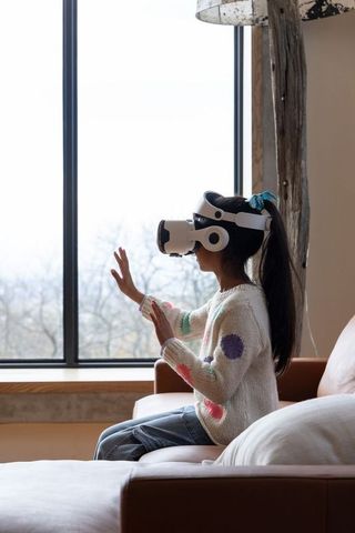 Asian girl exploring virtual reality on sofa wearing VR headset by window