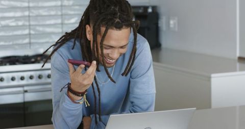 African american man working from home on laptop while leaving voice message on smartphone
