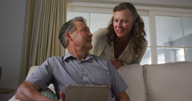 Mature couple enjoying a moment together in a cozy living room, woman leaning over the couch while man is seated and holding a tablet. Can be used for concepts of family, relaxation, technology use among seniors, and quality time. Ideal for websites and marketing materials focusing on senior lifestyle, tech-savvy older adults, or family-oriented products.