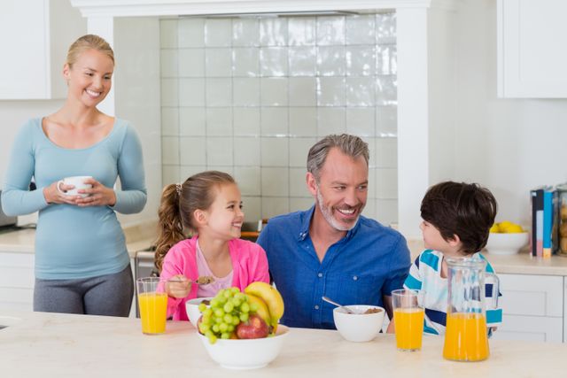 This image depicts a joyful family having breakfast together in a modern kitchen. The father and children are seated at the table, enjoying their meal, while the mother stands nearby with a cup. This image can be used for advertisements, family-oriented content, healthy lifestyle promotions, and articles about family bonding and morning routines.