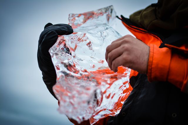 Person wearing gloves holds a clear, irregularly shaped ice block in an outdoor, winter setting. This image is ideal for use in articles, blogs, or advertisements related to cold weather activities, winter gear, climate studies, or nature exploration.