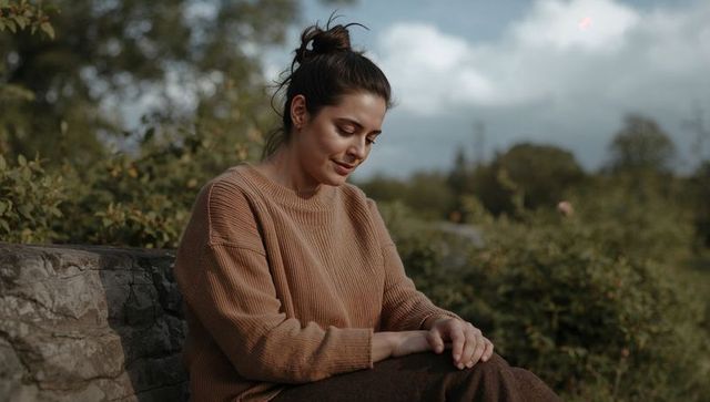 Contemplative woman sitting on stone wall in meadow wearing brown ribbed sweater under overcast sky