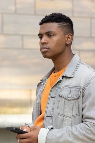 Thoughtful african american teen holding phone wearing denim jacket and orange sweater