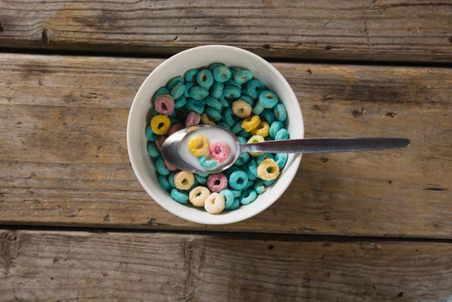 Colorful cereal in a white bowl with a spoon on a rustic wooden table. Ideal for breakfast-themed content, healthy eating promotions, or food blogs. Can be used in advertisements for cereal brands or morning routine articles.