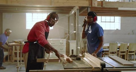 Carpenters cutting wood on table saw in busy workshop wearing safety gear