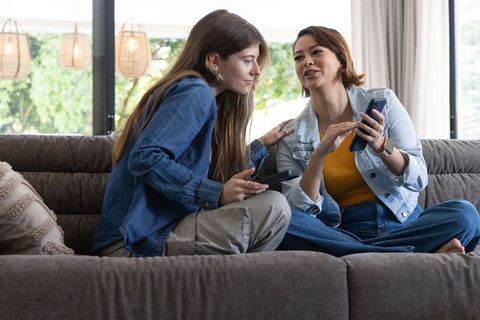 Friends sharing phone and remote on cozy living room sofa, chatting and laughing