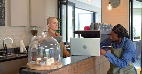 Diverse baristas collaborating behind counter using laptop and notebook in bright café