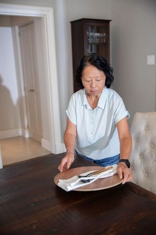 Senior chinese woman arranging tableware on dark wood dining table wearing smartwatch