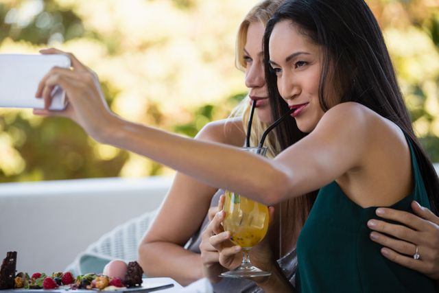 Two female friends are taking a selfie while enjoying drinks at an outdoor restaurant. They are smiling and appear to be having a good time. This image can be used for promoting social gatherings, restaurants, summer activities, and lifestyle blogs.