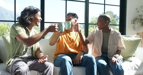 African american friends toasting with pastel mugs on sunlit sofa by black-framed window