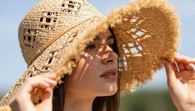 Woman wearing wide-brim straw hat holding brim casting patterned sun shadows on face