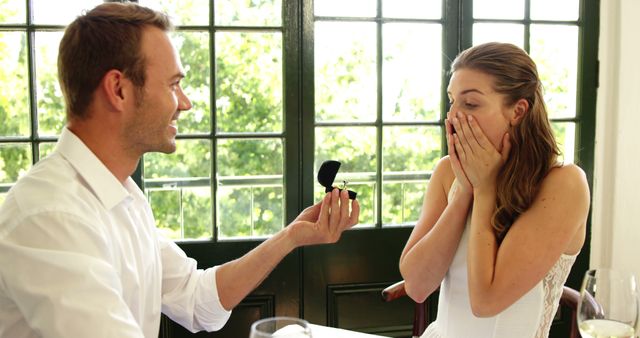 Smiling caucasian man proposing marriage with engagement ring to shocked woman in sunny restaurant. Marriage, engagement, ring, couple, relationship, tradition and lifestyle, unaltered.