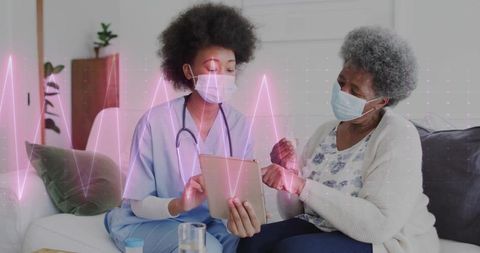 Nurse wearing mask pointing at tablet during home care consultation with senior woman