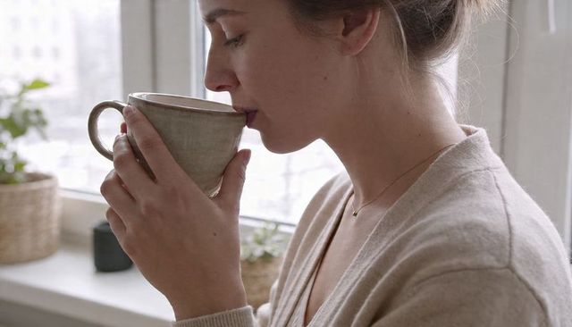 Sipping morning coffee by window in beige cardigan holding ceramic mug, cozy neutral scene