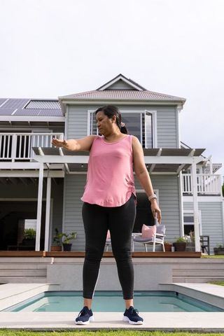 Mature woman standing at backyard plunge pool extending arm wearing pink top and leggings