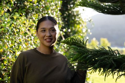 Asian woman standing in sunlit garden holding conifer branch, serene outdoor portrait