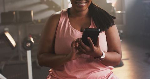 African american woman smiling and checking phone on gym bench wearing smartwatch