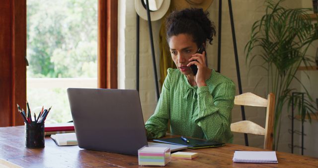 A professional woman working from her home office, multitasking between a phone call and using her laptop. She appears focused and engaged in her task. Useful for topics related to remote work, professional women, home office setups, and productivity.