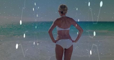 Standing woman wearing white bikini facing turquoise ocean on sunny beach
