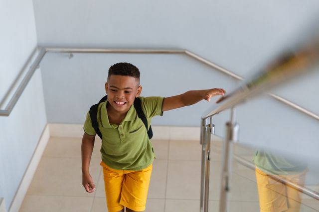 This image shows a cheerful African American elementary schoolboy climbing steps inside a school building. He is wearing a green shirt and yellow shorts, carrying a backpack, and smiling. This image can be used for educational materials, school promotions, childhood development articles, or back-to-school campaigns.