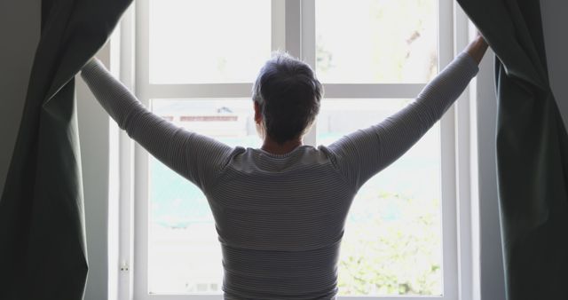 Rear view of senior caucasian woman at home drawing curtains at window in morning. Mental health, domestic life and senior lifestyle, unaltered.