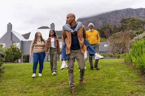 Multigenerational african american family walking on lawn with teen piggyback near metal-roof home