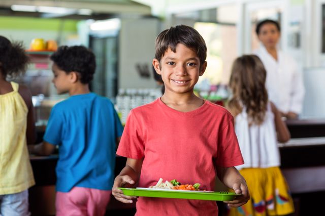 Schoolboy holding food tray in a school canteen, smiling at the camera. Other children are in the background, getting their lunch. Ideal for use in educational materials, healthy eating campaigns, and school-related content.