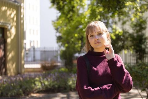 Young woman standing in sunlit urban courtyard wearing maroon sweater, contemplative student