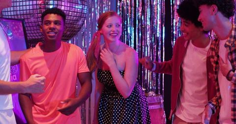 Laughing multiracial friends dancing and chatting under disco ball at retro neon party