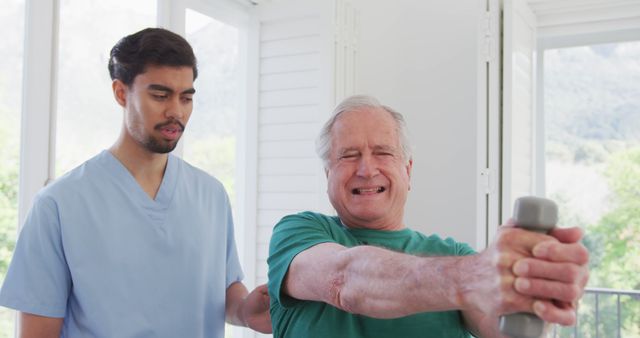 Senior man doing physical therapy with young male therapist in light-filled room. Older male holds small dumbbell while therapist provides assistance and guidance. Ideal for use in health and wellness advertisements, elder care promotions, and physiotherapy training materials.