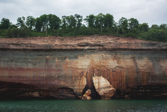 This striking nature scene displays a heavily eroded cliff face meeting a serene lake, topped with a lush and dense tree line. The colors and textures of the rock layers reflect the passage of time and elemental exposure. Ideal for use in nature documentaries, travel blogs, geological studies, or outdoor adventure promotion.