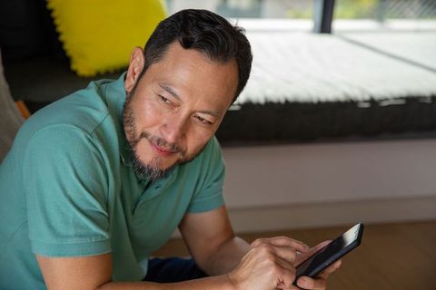 Mature asian man tapping smartphone in sunlit living room wearing teal polo and beard